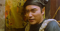 Close-up image of a person squinting in the rain, leaning against a wall with a yellow handkerchief on display that has handwritten black calligraphy on it. The person has ropes over their shoulders.