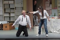 A young woman in jeans and sneakers holds a pistol to the temple of a man on his knees in a dress shirt and untied bowtie.