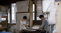 Two men work in the kitchen of a rice cake bakery. A work of seal style calligraphic work is framed, and blocking the window behind them.