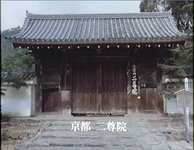 A large temple gate has a sign to the right of the door. It has the name of the temple on a calligraphic board. The name is also on a superimposed subtitle at the bottom of the frame.