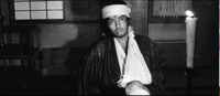 A bandaged man sits in front of a frame with black calligraphy, in black and white cinematography.