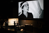 A photograph of the full stage space. One small screen on the left displays a hand projected from behind by overhead projector. Sarah Fornace, the actor playing Frankenstein, stands in front of this screen while another performer grabs her shoulder from behind. A second overhead projector casts the image of her frightened response onto a larger screen that dominates the stage, along with the hand projected onto the first screen.