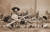 Black and white photograph of a woman posing with an oar in a dugout canoe. The canoe is full of vegetables, and the staged river is lined with vegetation.