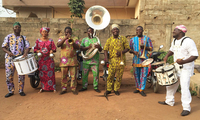 Seven band members, six men and one woman, pose for a photo in front of the brick concrete wall of a residential compound. Left to right, members are playing bass drum, shakers, flugelhorn, sousaphone, two trombones, and snare drum. A motorcycle can be seen in the background. Musicians are dressed in colorful African clothing.