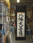 A photo of a tall vertical calligraphy scroll hanging in a storage area, black calligraphy on white paper, framed by black.
