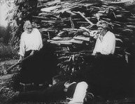 A woman holding an axe and a man stand in front of a pile of wood scraps. Between them, a man lies on the ground.