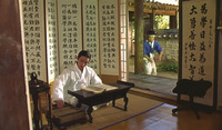 A film still of a man in white traditional clothes, seated at a small table, with calligraphy-covered screens behind and diagonally across from him. A man in blue and white approaches from the right, from outside.