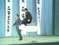 Color still of duo Konto 55-­gō on stage. Hagimoto Kin’ichi is pictured mid-­jump as his feet strike the back of partner Sakagami Jirō.