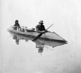 A black-and-white photograph of a couple in a canoe. Several boxes and packages are being transported in the canoe as well.