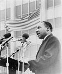Black and white image of Rev. Martin Luther King Jr. speaking at a podium with an American flag waving in the background.