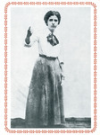 Full-length photo of Flynn speaking at a rally. Her long hair is pulled back. One arm emphasizes her words. She wears a long skirt and a blouse with a scarf.