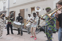 A street performance with an eight-­member ensemble including (from left to right) tenor saxophone, flugelhorn, bass drum, snare drum, soprano saxophone, two trombones (one is the author), and, in the rear, sousaphone. Performers wear custom-­made African clothing. A small stage platform and a tall wooden drum can be seen in the background.