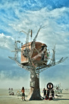 Photograph of Five Ton Crane’s Steampunk Treehouse. An approximately 25-­foot tall sculpture of a tree with treehouse atop it. Tree trunk and house are rusty metal; the tree’s limbs are stainless-­steel crane-­like armatures reaching skyward, with various objects hanging from them.