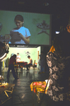 Photograph depicting a female actor being filmed onstage and projected onto a screen above the set, with lighting unit and videographer in the foreground.