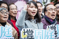A young woman, Agnes Chow Ting, stands surrounded by a group of people chanting at a rally. Her right arm is raised, bent at the elbow, and she is making a fist. Several people hold signs with Chinese text