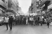 A wide group of Israeli Black Panther demonstrators of various ages with Hebrew protest signs march together across a city street.