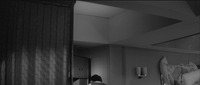 Another unusual view of Misako’s apartment, with the patterned ceiling dominating the top of the frame, at an angle, while the space below is bisected by a moveable partition wall blocking the Left side of frame. At the bottom of the screen, just Left of center, is the back of Hanada’s head and shoulders as he cradles the phone in his neck. On the Right we glimpse the pile of stacked-­up beddings and furniture.