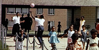 Color photograph of Black children playing ball of various ages and genders, with other children playing in front of them, facing the camera.