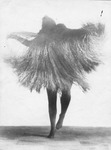 Photograph of Mary Wigman dancing solo in a straw costume, which obscures her body and makes her appear like a spirit with legs in tights. One leg is stepping up with the other lightly on the floor; only one arm is visible within the straw and is positioned away from her body.