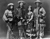 Actors Ned Buntline, William F. Cody, Mlle. Morlacchi, and Texas Jack Omohundro stand in pose, the men wearing fringed buckskin and holding rifles, Mlle. Morlacchi wearing a fancy dress with bead trim.