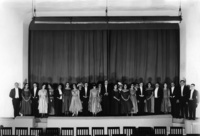 In this photograph, the cast of Princess Turandot stands on the forestage in evening dress, the curtain closed behind them, looking directly at the audience. Four commedia characters, their faces painted to look like masks, peer between them.