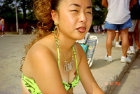 Lee sits outside on the curb wearing a yellow flowered bikini top, a nameplate necklace in gold, and large gold hoop earrings. Her lipstick is pale pink with dark liner. She is slightly tanned.