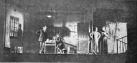 Production photograph depicting four actors in a set of a semiunderground apartment, with heavy shadows on the walls.