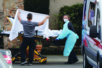 A young white woman with her hair pulled back into a ponytail wears protective gear as she pulls a stretcher toward a hospital vehicle. A young man in a grey t-shirt is seen from behind as he holds a white sheet to conceal the COVID-19 patient being moved on a yellow stretcher.