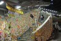 A young woman climbs an outdoor staircase that is completely covered with Post-It notes bearing messages from activists. A banner hung from the ceiling reads, “Lennon Wall Hong Kong” while a sign over the staircase says, “We are not alone