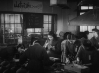 Women work in a factory beneath a banner on the wall and a poster in the window with black and white calligraphy, in black and white cinematography.