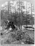 A black and white photograph of three men lounging beneath a makeshift canvas tent on the shore with the river flowing behind them. Their birch-bark canoe sits beside them propped up agains a pine tree