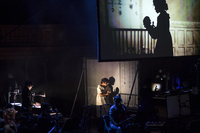 Photograph of the actress playing Mary Shelley standing onstage holding her infant daughter, with multiple projections of her shadow displayed behind her. The stage image uses the same multiple screen and projection strategies as in the previous photo.