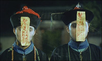 A film still of two individuals standing side-by-side in period clothing, with face-covering paper hanging from their hats, covered in red calligraphic text.