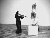 Figure 9. Violinist Mari Kimura, wearing a formal black dress, stands near Guitarbot, a four-stringed robotic instrument on a pedestal that is considerably taller than she is, and looks at it as if it were a fellow musician.