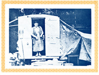 Carr stands at the door of her trailer, smiling; she wears a long dress of printed fabric and a vest. The trailer has a tent-like area on the right; a broom hangs to the left.