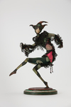 The photograph depicts a wax doll by Lotte Pritzel from circa 1917, called Harlequin. Masked and dressed in a green and pink diamond-patterned costume adorned with lace and frills, the dancing Harlequin, who stands on pointe on one leg, knees and arms bent, is shown frozen in a jumping move, the frills of his costume mid-flutter.