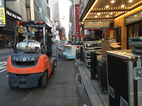 View of city street, black boxes with steel reinforcement lined up on the sidewalk under a lit theater marquee. Scenic element in the distance of diner stools and counter.