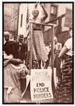 A Black woman on a soapbox, arm raised, speaks to a crowd of men, women, and children, Black and white, rallying against police shootings. American flag beside speaker.