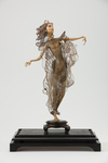 The photograph depicts a wax doll by Lotte Pritzel from 1923, called Female Dancer/Bayadere. The elongated, androgynous-looking female figure standing on a plinth is wrapped from head to toe in see-through gauze and lace with a golden trim, which uncovers her entire body torso, revealing her breasts, abdomen, and pubis. With undulating arms and wings in the back, the doll resembles a dragonfly ready to take flight.