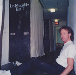 Man sitting on a floor with legs stretched out in a hallway; a tall, black road-box labeled “Les Misérables: Tour 1” stands on the opposite side of the hallway.