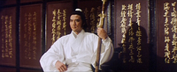 A man in white holds a sword, sitting in front of several wall plaques with gold calligraphy.