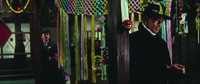 Colorful, theatrical evocation of a city marketplace emphasizing the Man in Black (hat and coat) Right of screen, looking down, next to the wooden beam (Right of center) of the little shop behind him. The beam dominates the foreground, decorated with hanging fruit, little signs, and other paraphernalia. Middle ground center, the shop entrance is decorated with dozens of yellow, pink, and blue streaming ribbons hanging from decorated latticed work similarly colored. Background Left, a woman in profile and a plainclothesman, watching the outlaw, are seen in the space between door and display.