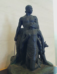 This is a photo taken from the right side of a bronze sculptural group, two-­thirds life size, placed on a pedestal. The central figure is a young, muscular Japanese male nude, his sleek hair tied into a knot at the nape of the neck. He looks forward heroically, holding a sword in his right hand while helping another man onto his knees with his left. His right foot points back, and his left leg extends forward. The kneeling Asian man is only wearing a loincloth. His body is emaciated, his left hand resting on his chest while looking up at the young Japanese with sadness and gratitude. Partially visible are a hunting dog and a young boy standing to the left of the Japanese young man, as well as a woman hiding behind his back, sheltered by him.