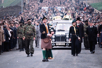 In a large funeral procession, hundreds of people walk behind a hearse, bagpipers, and soldiers dressed in camouflage.