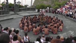 Group of about 30 men wearing checkered sarongs perform in a circle on a circular stage, surrounded by tourists.