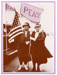Marching on a street wearing dresses, hats, and white gloves, Addams holds a large peace flag and McDowell holds an American flag. Car in background with peace flag.