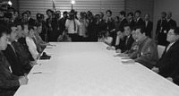 A large table with formally dressed people on the right and left sides. There are a few papers and notebooks in front of them. Photographers and onlookers line the far side of the table.