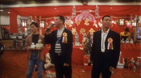 Three people, two wearing jackets and colorful ribbons, stand in front of a red stall. Calligraphy is visible in the background.
