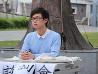 A college student, Wong Ching-Fung, sits at an outdoor table. Photographed from the waist up, his arms rest on the table and his hands are folded. He is looking past the camera into the distance. Paper signs with Chinese text hang from the front of the table
