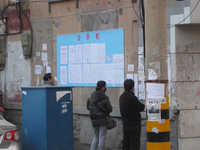 Photograph of Chinese notices pasted onto a public notice board attached to a dirty wall, with three people attentively reading through the announcements.